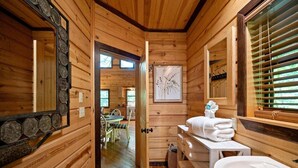 Ein Holzblockhaus-Interieur mit Holzboden, einem Essbereich inklusive Tisch und Stühlen, einem weißen Waschbecken und einem Spiegel mit Zierrahmen.