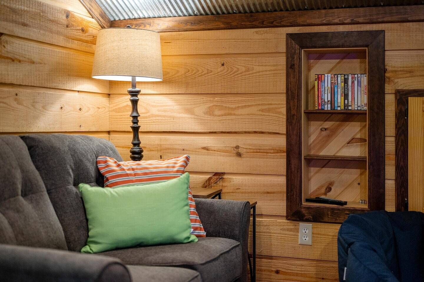 A cozy room with a wooden log wall, a grey sofa with green and striped pillows, a floor lamp, and a bookshelf filled with books.
