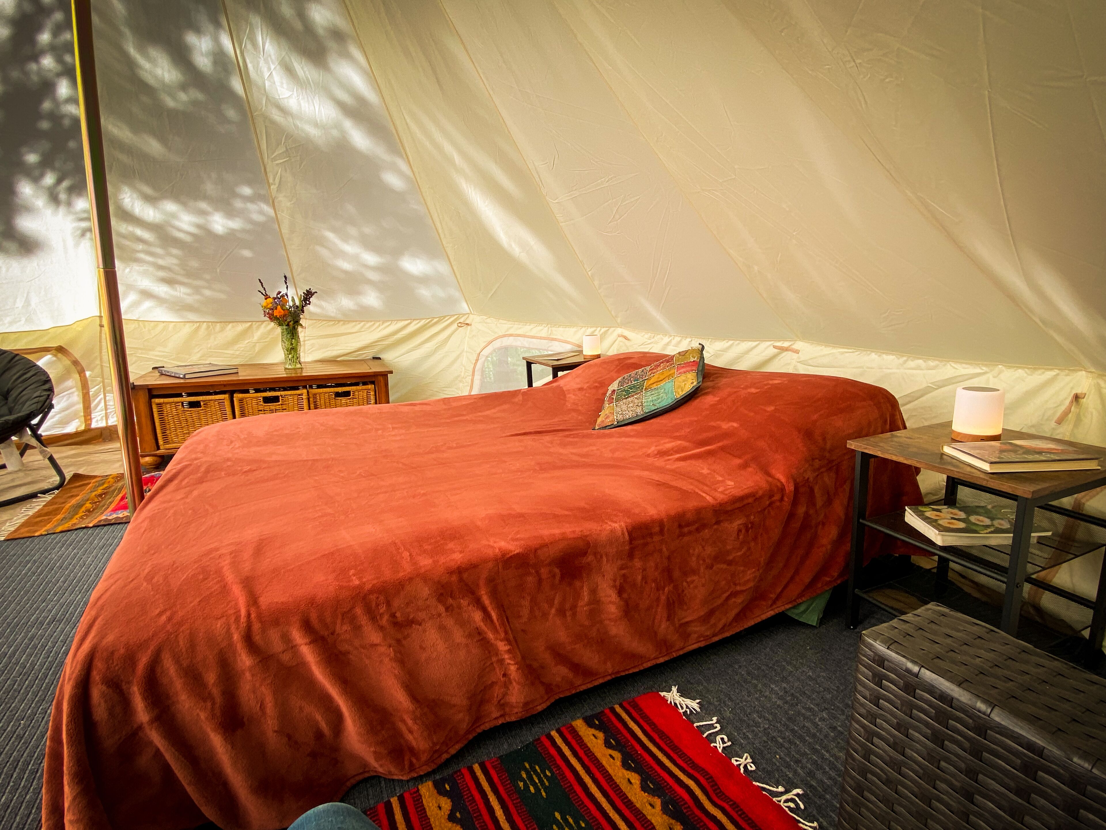 Een tentruimte met een groot bed, een klein tafeltje met een lamp en een rieten mand.