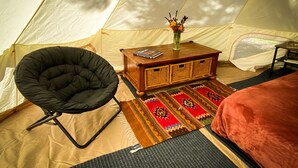 L’intérieur d’une tente, avec une chaise noire, une table d’appoint en bois sur laquelle se trouvent un vase et des livres, un tapis à motifs et un lit recouvert d’une couverture rouge.