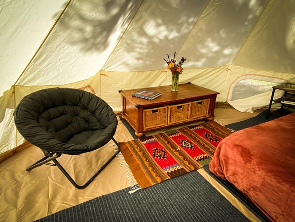 A tent interior with a black chair, a wooden side table with a vase and books, a patterned rug, and a bed with a red blanket.