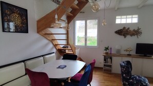 A dining area with a wooden staircase, a white table with chairs, and a television on a wooden cabinet. - Charming little house 5 minutes' walk from the beach (Locquirec)