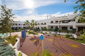 A playground with swings and a climbing structure, surrounded by a landscaped area with palm trees and a pool.
