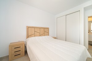 A bedroom with a wooden headboard, a bed with a white bedspread, a nightstand with drawers, and a closet with sliding doors.