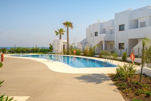 A modern pool area with a wooden deck, surrounded by white buildings and palm trees.
