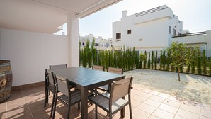 An outdoor patio with a table and chairs, a barrel, and a view of modern buildings and a garden. - Apartamento Babilonia (Mar de Pulpi) (San Juan de los terreros)