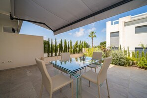 Eine Terrasse im Freien mit einem Glastisch und vier Stühlen, umgeben von einem modernen Gebäude und verschiedenen Pflanzen.