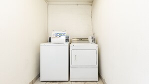 A laundry area with two white appliances, one larger and one smaller, in a corner with white walls and a tiled floor.