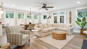 A spacious living room with a large sectional sofa, a dining area with a table and chairs, and a ceiling fan. - High-End Beach Home, Walking Distance to Beach & Close To Town - On The Half Shell! (Folly Beach)