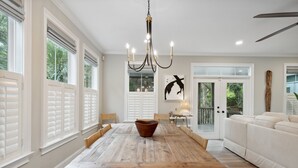 A spacious dining area with a wooden table and chairs, a chandelier, and a view of the outdoors through large windows. - High-End Beach Home, Walking Distance to Beach & Close To Town - On The Half Shell! (Folly Beach)