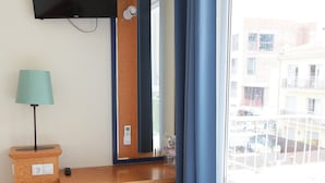 Quarto de hotel com cama, escrivaninha, cadeira, televisão e janela com vista para edifícios.
