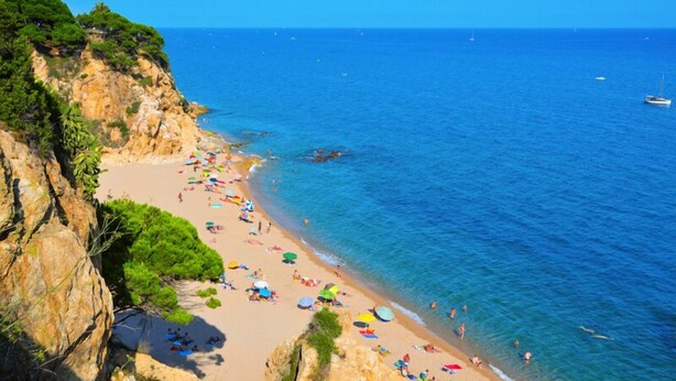 Uma praia com águas azul-claras, falésias rochosas e pessoas aproveitando o sol.