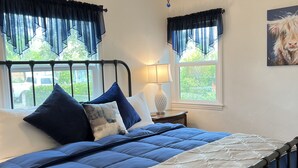 Een slaapkamer met een bed, een plafondventilator, een raam met gordijnen, een lamp en een schilderij aan de muur.