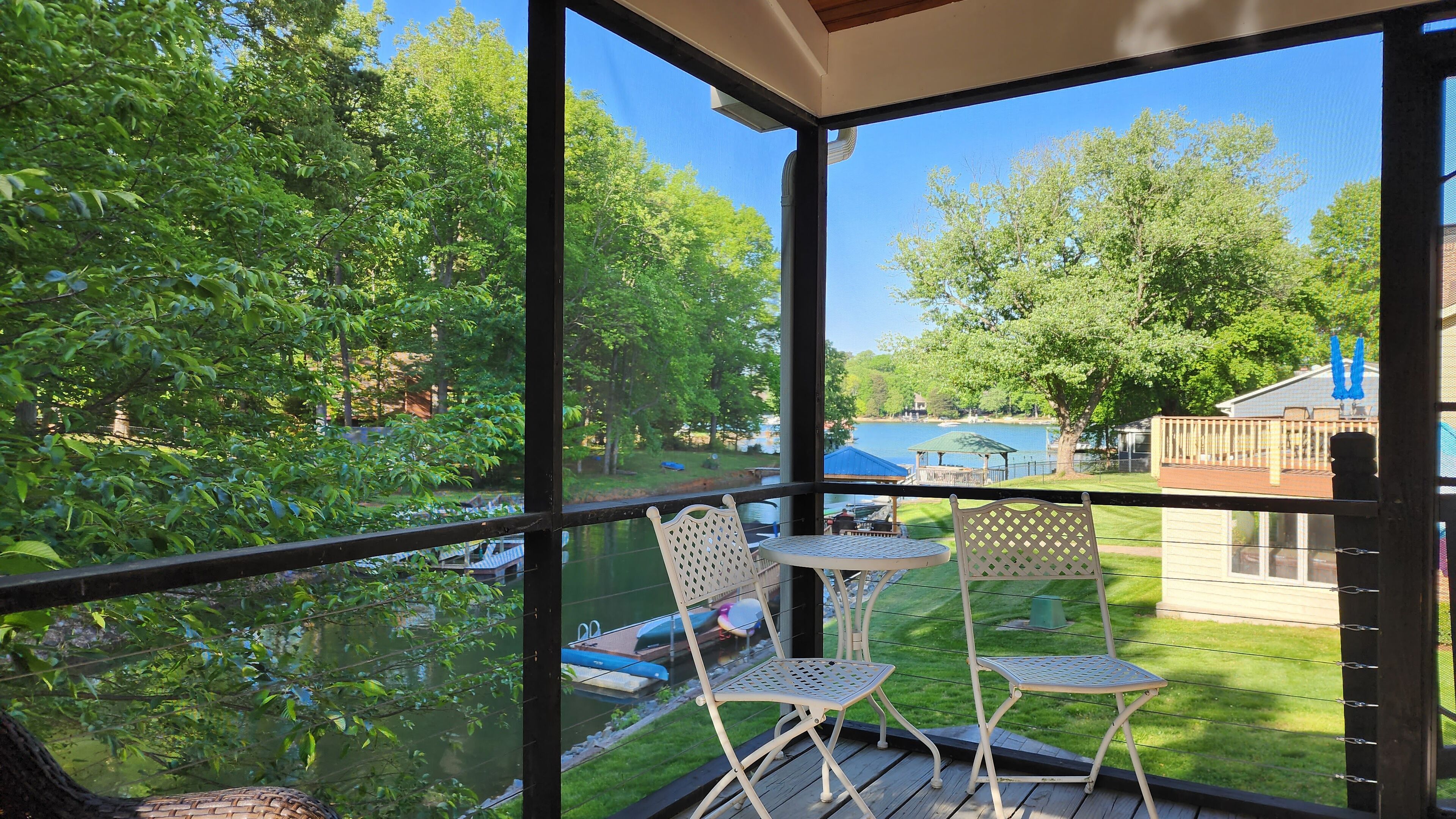 Un espacio exterior cubierto con dos sillas metálicas y una pequeña mesa, con vistas a un lago y una zona verde con árboles.
