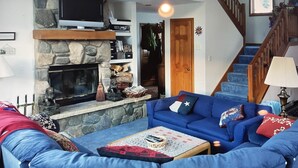 Una sala de estar con chimenea de piedra, un sofá azul, un televisor y una escalera.