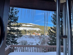 A view from a balcony with a wooden railing, looking out at a snowy landscape with trees and a mountain in the background.