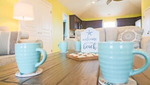 Un salon chaleureux avec des tasses turquoise posées sur une table en bois, un panneau indiquant « Bienvenue à la plage » et un ventilateur de plafond.