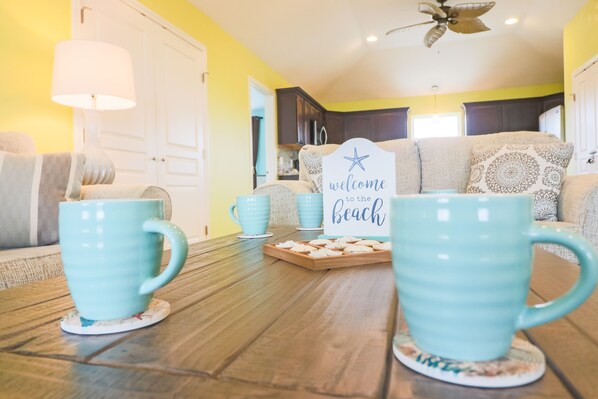Ein gemütliches Wohnzimmer mit einem Holztisch, zwei türkisfarbenen Tassen, einem Schild mit der Aufschrift 'welcome to the beach' und einer Couch mit gemusterten Kissen.