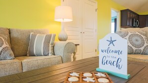 Un salon avec un canapé, une table basse en bois sur laquelle sont disposés des biscuits en forme d’étoiles de mer, une lampe et une pancarte indiquant « Bienvenue à la plage ».