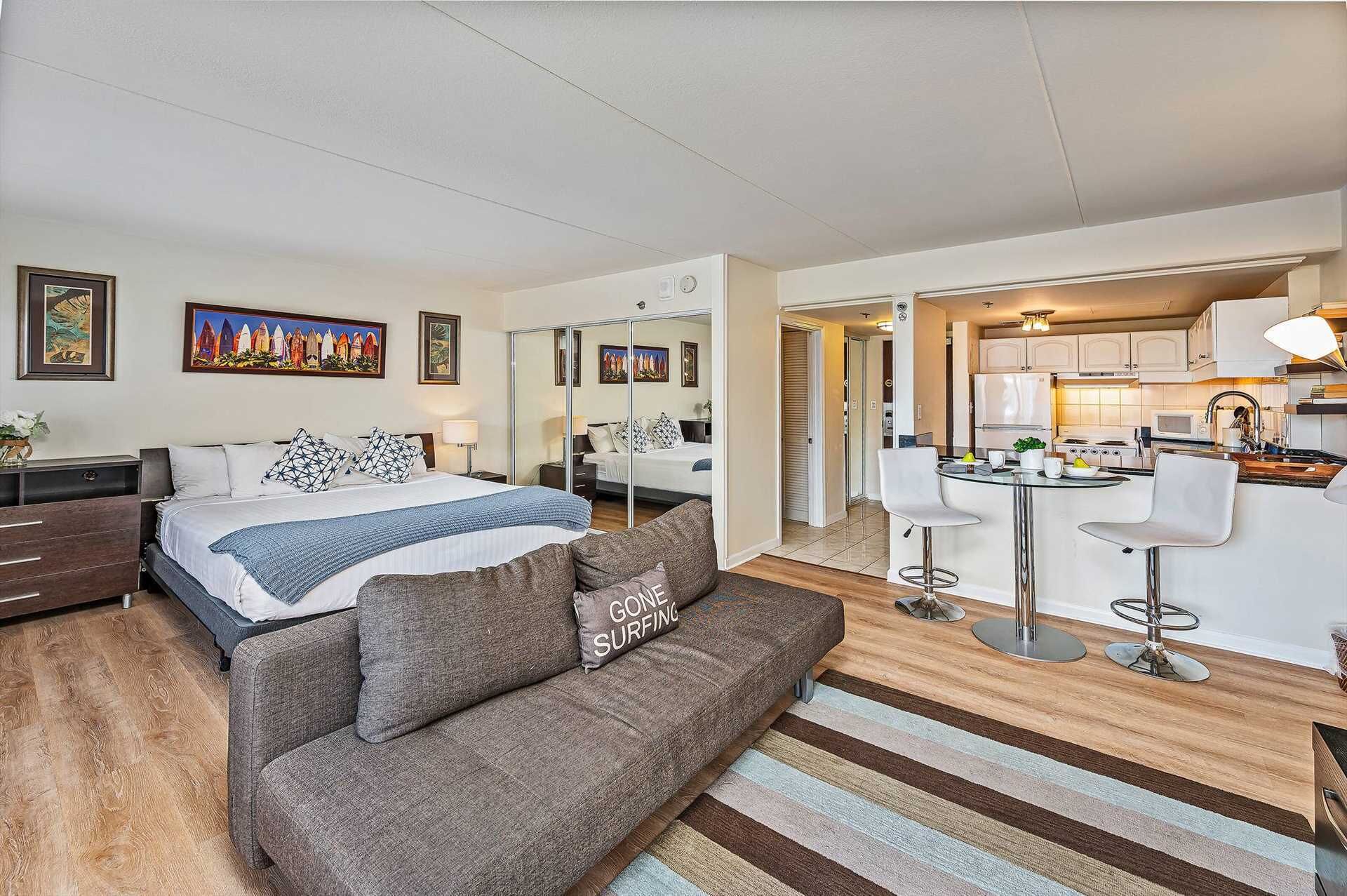 A modern hotel room with a bed, a sofa, a small kitchen area, and a dining space.