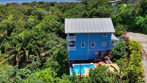 A two-story house with a blue exterior, surrounded by lush greenery and a pool. - Sunset and Ocean views at Sandy Feet Retreat with private infinity pool. (Sandy Bay)