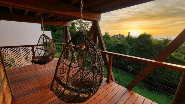 A wooden deck with hanging woven chairs and a view of a sunset over a forest. - Sunset and Ocean views at Sandy Feet Retreat with private infinity pool. (Sandy Bay)