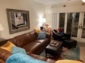 Un salon avec un canapé en cuir marron, une table basse et un fauteuil. Un tableau encadré est accroché au mur, il y a une lampe et une fenêtre avec des rideaux.