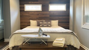 Un lit bien fait, avec une tête de lit en bois, deux tables de chevet sur lesquelles se trouvent un fer à repasser et des serviettes, et deux fenêtres équipées de stores.