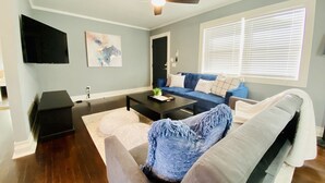 Un salon avec un canapé bleu, un divan blanc, une table basse, un ventilateur de plafond et une fenêtre avec des stores.