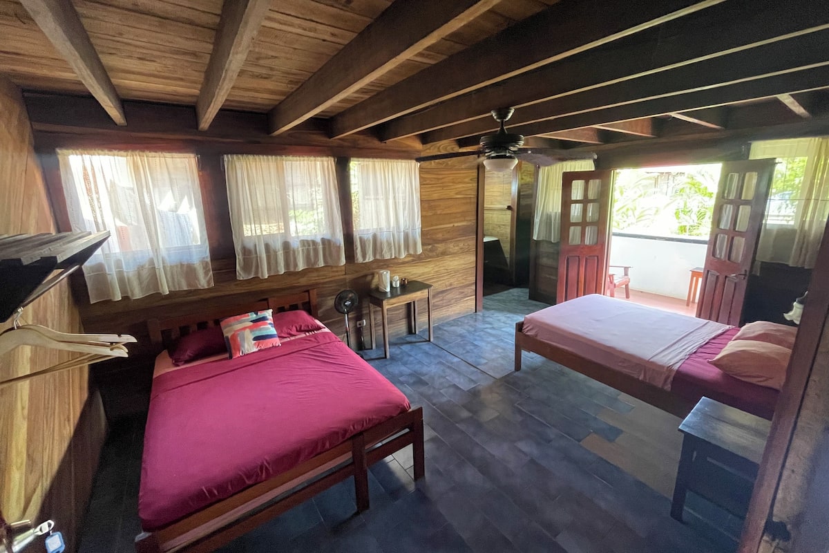 A room with two beds, wooden ceiling beams, a ceiling fan, and a window with curtains.