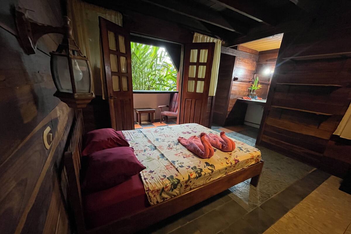 A wooden room with a bed, a small table, and a window with a view of greenery.