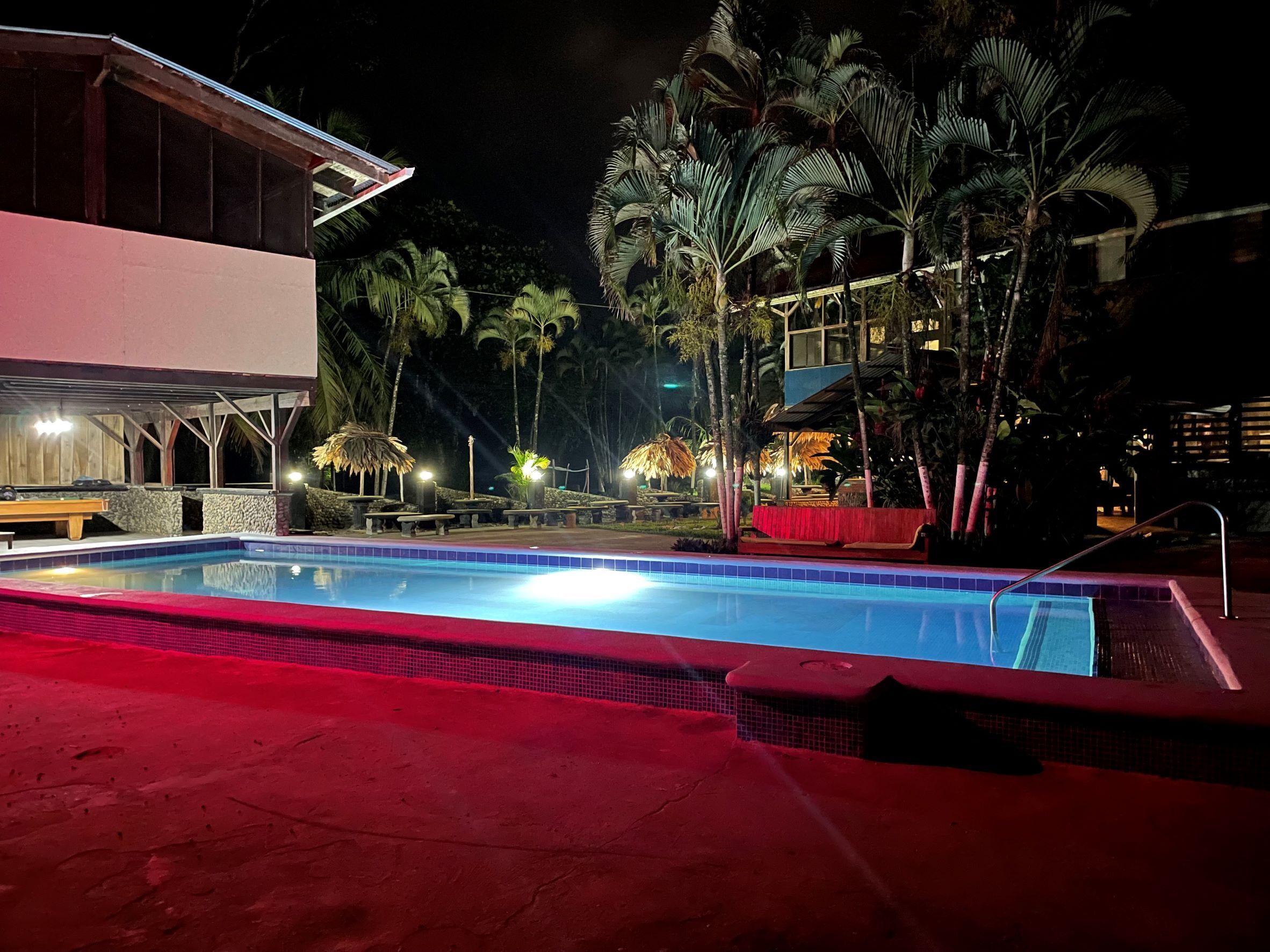 A nighttime pool area with a red-tiled pool, lounge chairs, and palm trees.