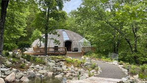 A rustic cabin with a stone exterior, surrounded by a natural landscape with a pond and wooden deck. - Unique Dome Home With Hot Tub & Piano, on 28 acres (New Paltz)