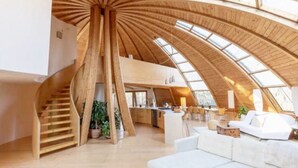 A spacious loft with a high ceiling and skylights, featuring a central staircase, a kitchen area, and a living space with a white sofa. - Unique Dome Home With Hot Tub & Piano, on 28 acres (New Paltz)