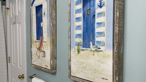 Een hal met twee wanddecoraties van kustlandschappen, een witte deur en twee witte handdoeken op een handdoekrek.