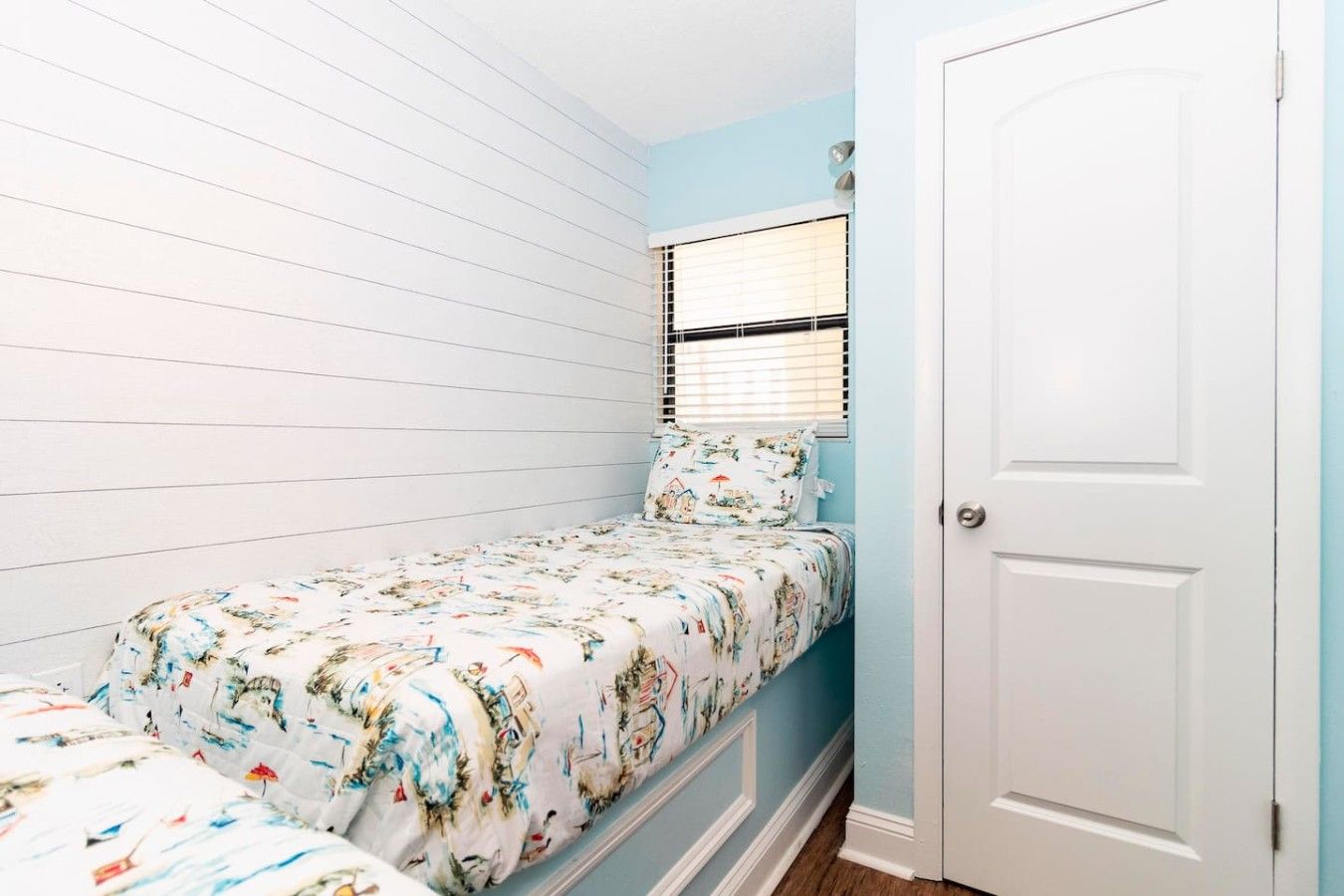 A small bedroom with a single bed, a window with blinds, and a white door.