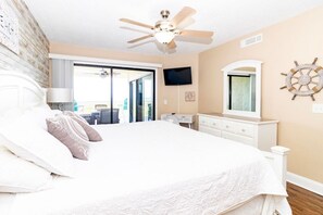 A bedroom with a bed, a desk, a television, a ceiling fan, and a view of the outdoors.