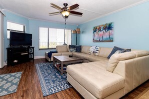 A living room with a sectional sofa, a coffee table, a TV on a stand, and a ceiling fan.
