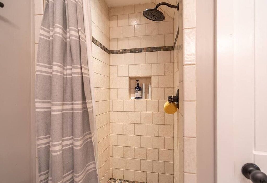 A shower with beige tiles, a showerhead, and a bottle of shampoo on the wall.