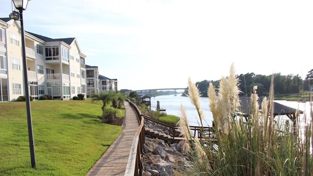 A waterfront property with a wooden boardwalk, a grassy lawn, and a view of a river and bridge. - Spacious condominium close to everything North Myrtle has to offer (North Myrtle Beach)