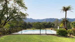A swimming pool surrounded by a black metal fence, with a grassy lawn in the foreground and mountains in the background. - Never Never Cottage (Gleniffer)