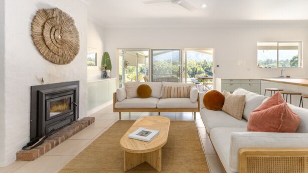 A modern living room with a fireplace, a sofa, a coffee table, and a large window offering a view of greenery. - Never Never Cottage (Gleniffer)