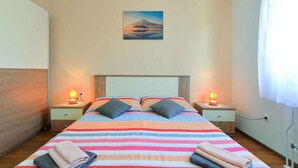 Een net opgemaakt bed met streepjesbeddengoed, twee bedlampen, een nachtkastje met een lamp en een schilderij van een berg aan de muur.