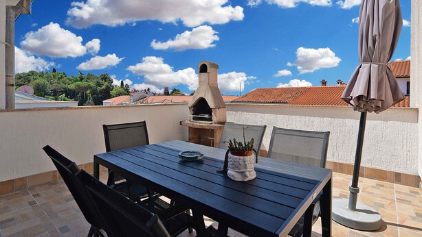 Een terras buiten met een tafel, stoelen, een barbecue en een parasol.