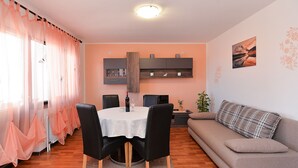 Een eetgedeelte met een tafel, stoelen en een bank. Er is een raam met dunne gordijnen en een wandplank met decoratieve objecten.