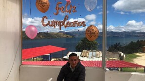 A man sitting in a room with a large window overlooking a scenic landscape with red-roofed buildings and a body of water. - Playa Blanca El mana (Boyacá)