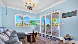 A spacious living room with a large sectional sofa, a wooden coffee table, and a view of a pool and palm trees through large windows. - P24 - Fabulous 4 bed, 4 bath home with pool and dock (Key Colony Beach)