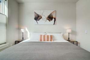 A bedroom with a large bed, two bedside tables with lamps, a wall-mounted artwork of elk, and a window with blinds.