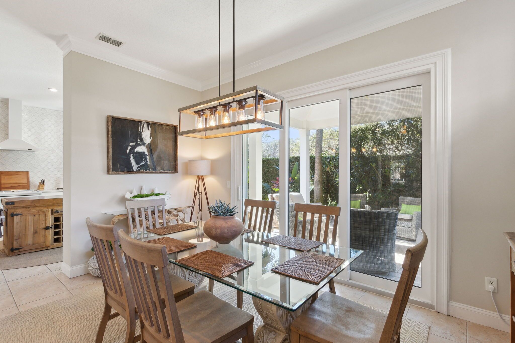 A dining area with a glass table, wooden chairs, a hanging light fixture, and a view through large glass doors to an outdoor patio.