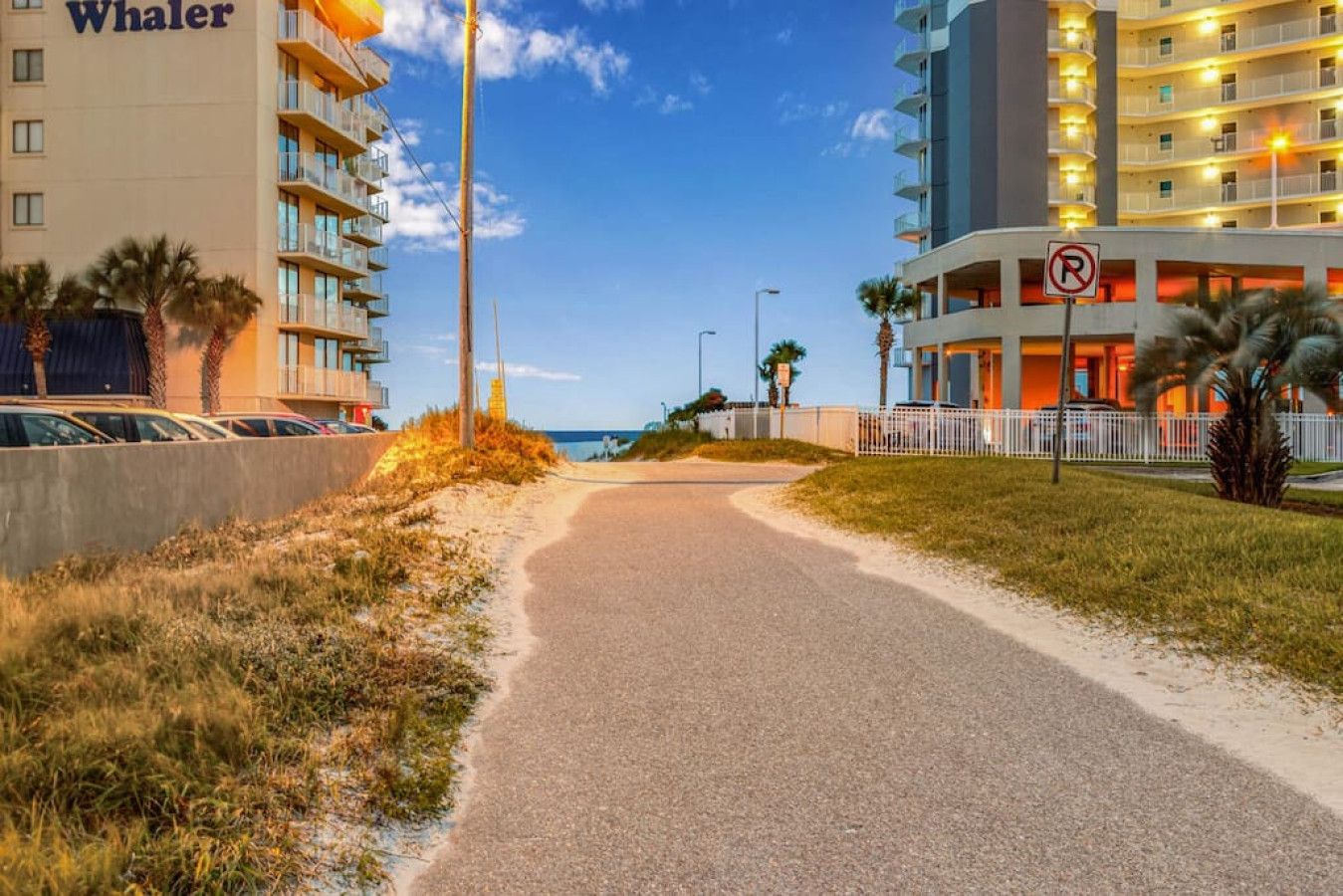 A road leading to a hotel with a 'No Parking' sign, a beach in the background, and a clear sky.